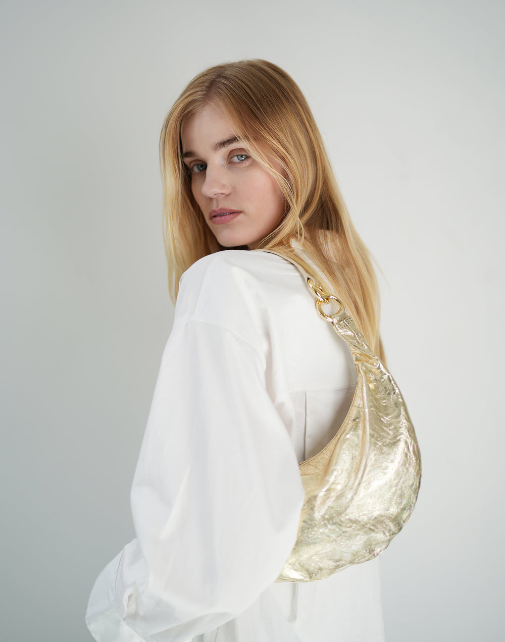a blond-haired woman wearing a white oversized buttondown and a gold leather crescent shoulder bag #color_gold
