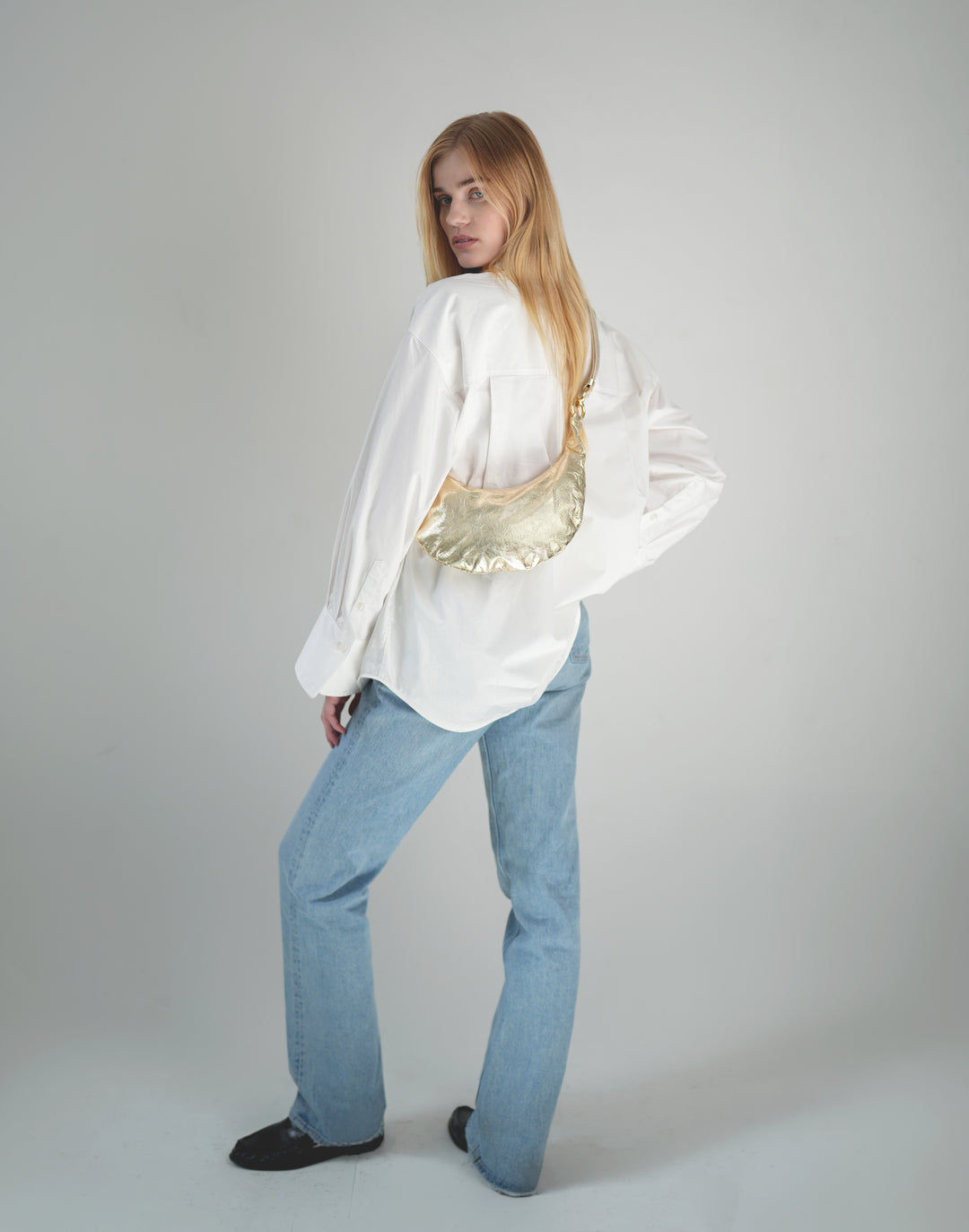 a woman stands with her back to the camera, wearing an oversized white button down, light blue jeans and black loafers and a gold leather crossbody crescent bag #color_gold