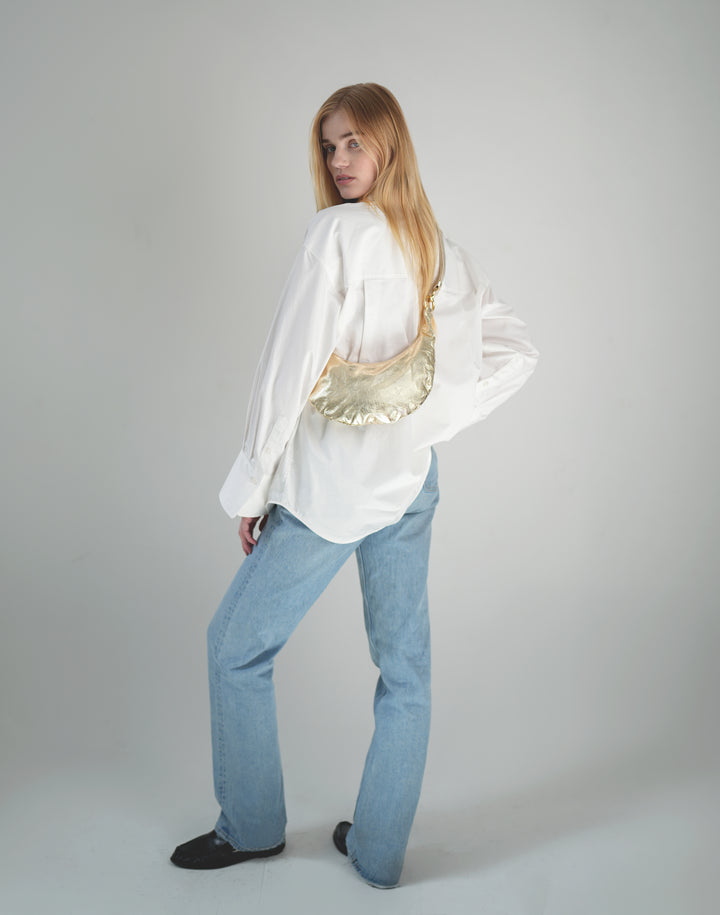 a woman stands with her back to the camera, wearing an oversized white button down, light blue jeans and black loafers and a gold leather crossbody crescent bag #color_gold