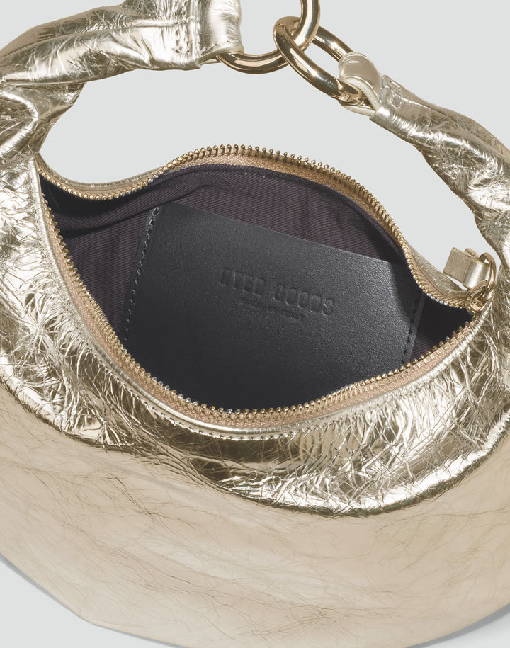 Close-up of a metallic gold textured handbag interior with a black leather insert and gold zipper, showing the brand name 'HYER GOODS' embossed on the pocket. #color_gold