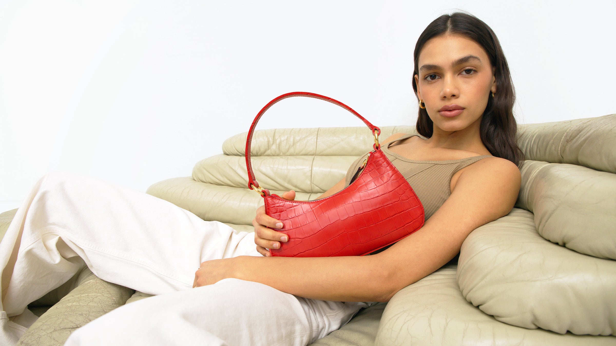 woman lounging on a beige vintage chair wears a tan top, white pants and a bright red croc mini shoulder bag.