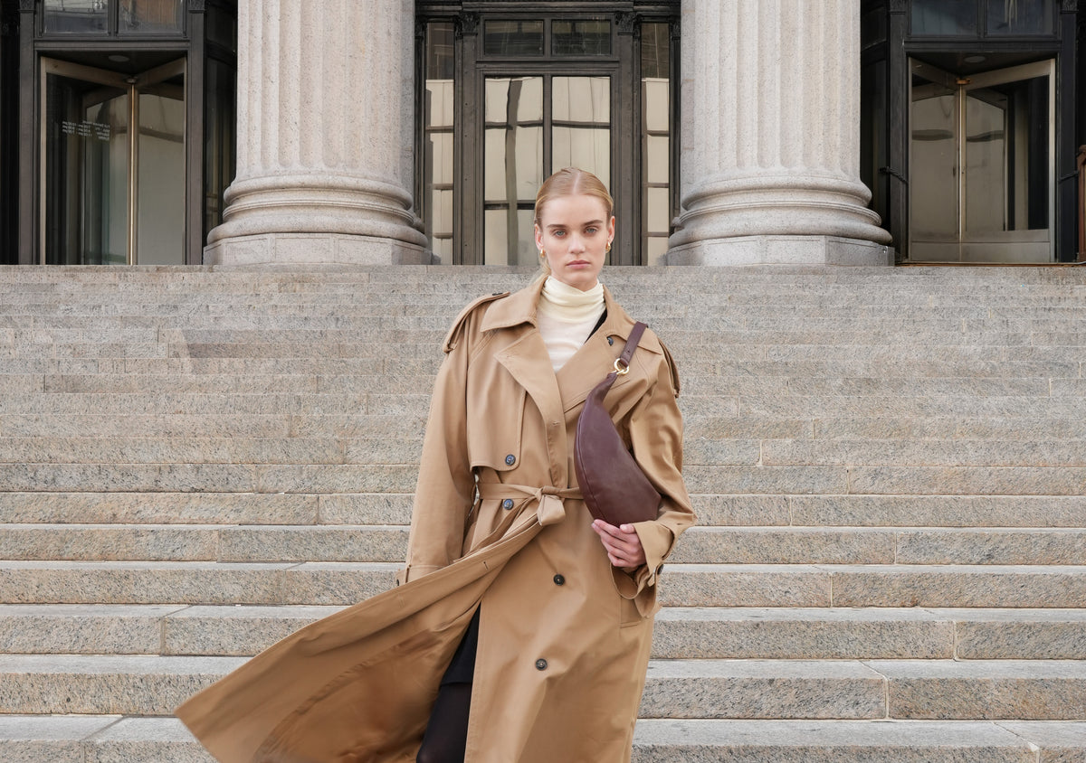 a woman stands on the steps of a regal-looking building, with tall columns behind her, wearing a cream turtleneck, trench blowing in the wind, and a HYER GOODS bordeaux vintage ring bag on her shoulder