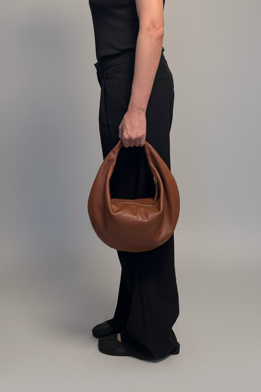A person wearing black clothing holds a large, slouchy brown leather shoulder bag in front of a brown wood cabinet and a light gray wall. #color_cognac